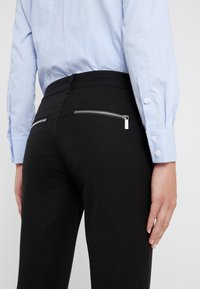 Pantalones negros ajustados con dos bolsillos traseros con cremallera, hechos de una tela suave. Llevados con una camisa de manga larga a rayas azul claro.