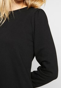 Top negro de manga larga con cuello redondo. Presenta detalles de hombros fruncidos y una tela suave y texturizada.