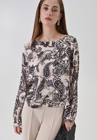 Blusa in bianco e nero con motivo paisley, realizzata in tessuto liscio, con maniche lunghe e ampia scollatura a barca, abbinata a pantaloni beige.