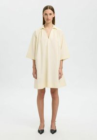 Vestido de manga corta con escote en V en tejido amarillo claro. Corte holgado, cae por encima de la rodilla con una textura suave. Combinado con tacones negros de punta.