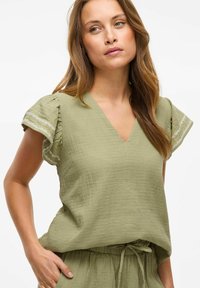 Blusa de color verde oliva, con textura, y escote en V con mangas con volantes. Presenta un sutil bordado blanco en los bordes de las mangas y un corte holgado.