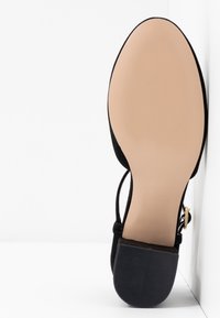 Chaussure plate noire avec une semelle beige lisse et un talon carré noir, vue de côté par dessous sur fond blanc.
