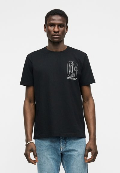 Νεαρός άνδρας φοράει μαύρο μπλουζάκι Off-White™ με λευκό λογότυπο στο στήθος και ανοιχτόχρωμο μπλε τζιν, στέκεται μπροστά σε απλό φόντο.