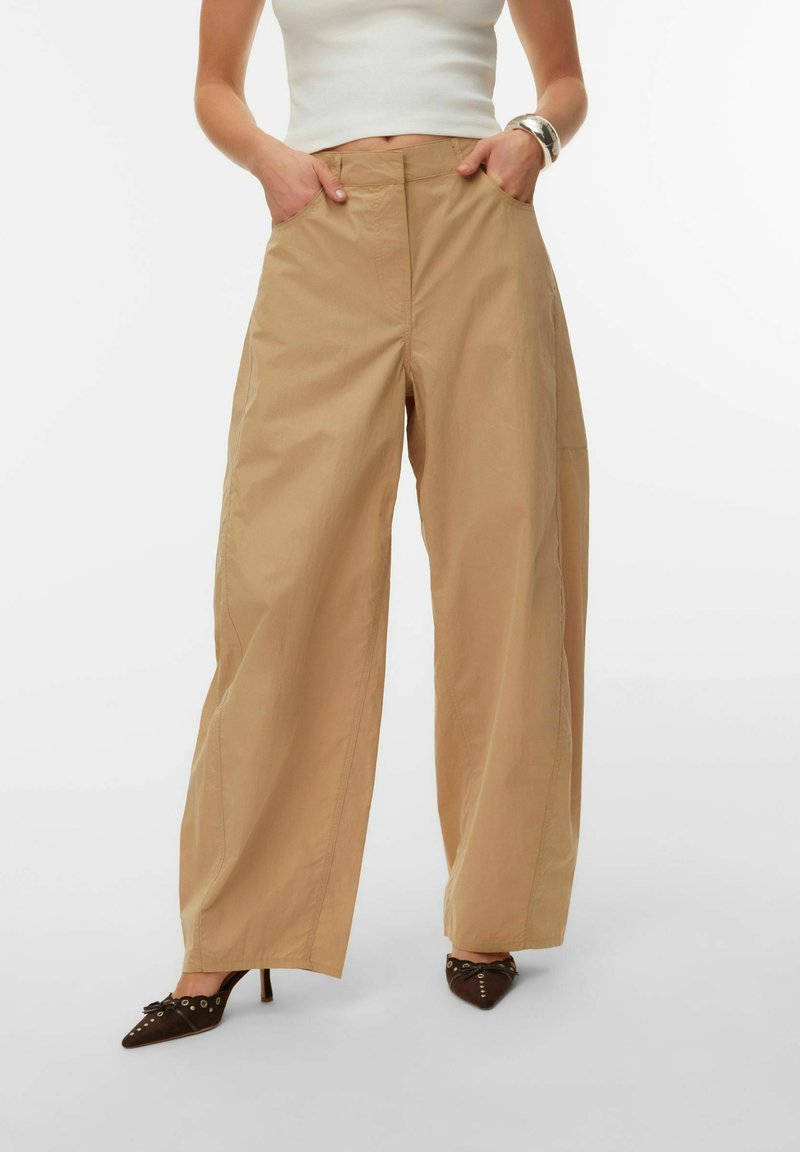 Femme portant un pantalon beige à jambes larges et un crop top blanc, les mains dans les poches, associée à des chaussures à talons hauts pointues marron.