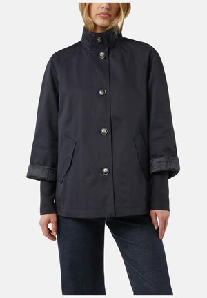 Kobieta w granatowej kurtce na guziki z wysokim kołnierzem, kieszeniami z patkami i mankietami na rękawach, ubrana w ciemne dżinsy, stojąca na jednolitym tle.