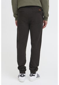 Donkerbruine joggingbroek met een elastische tailleband, voorzien van twee achterzakken en aansluitende enkels. Gecombineerd met witte sneakers.