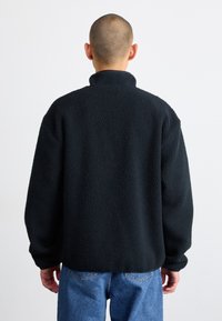 Pullover in pile nero con colletto alto e superficie testurizzata, abbinato a jeans azzurri chiari. Il design presenta una vestibilità rilassata e maniche lunghe.