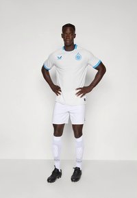 Bílý fotbalový dres s modrými akcenty, krátké rukávy, kulatý výstřih a kontrastní lemování na rukávech. Černé kopačky a bílé ponožky.