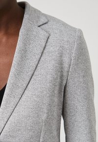 Pogledu od blizu na sivo teksturirano blejzer na osebi s temno kožo, ki prikazuje rever, ramo in zgornji del prsnega koša nad črnim topom.