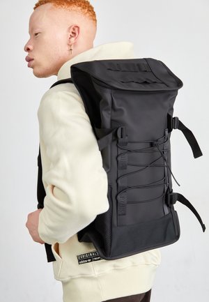 Person with short red hair wears cream hoodie and large black roll-top backpack with side straps and front bungee cords.