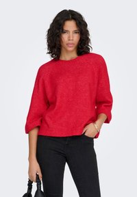 Pull-over rouge tricoté avec un col rond et des manches trois-quarts. Texture douce avec une coupe décontractée, associé à un jean noir.