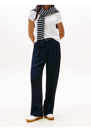 Biała koszulka z logo, granatowe spodnie o szerokich nogawkach oraz granatowo-biały pasiasty szalik przewieszony przez ramiona. Białe trampki z brązowymi podeszwami.