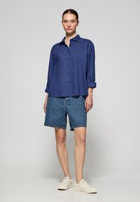 Marinfärgad skjorta med rullade ärmar, kombinerad med blå denimshorts. Modell bär ljusa sneakers och står mot en neutral bakgrund.