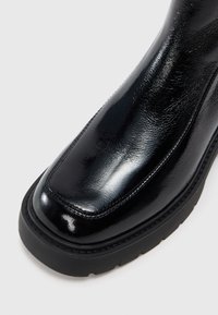 Crne sjajne gležnjače s okruglim nosem, glatkom kožnom teksturom i debelim crnim gumenim đonom. Ima minimalne šavove kao dekoraciju.