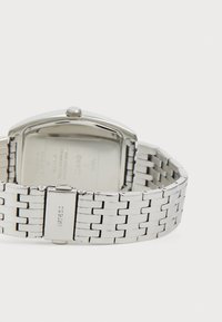 Bracelet de montre en métal argenté avec une fermeture gravée "GUESS", montrant l'arrière du boîtier de la montre et la couronne sur un fond blanc.