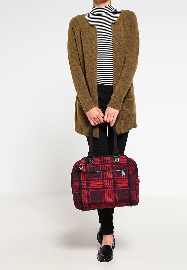 Personne portant un cardigan marron et un pantalon noir tenant un sac à main à carreaux rouge et noir avec des poignées noires.