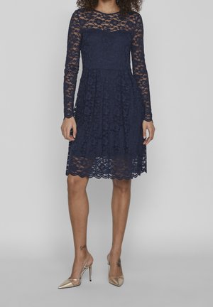 Vrouw in een marineblauwe kanten jurk tot op de knie met lange mouwen en beige hoge hakken, staand tegen een effen achtergrond.