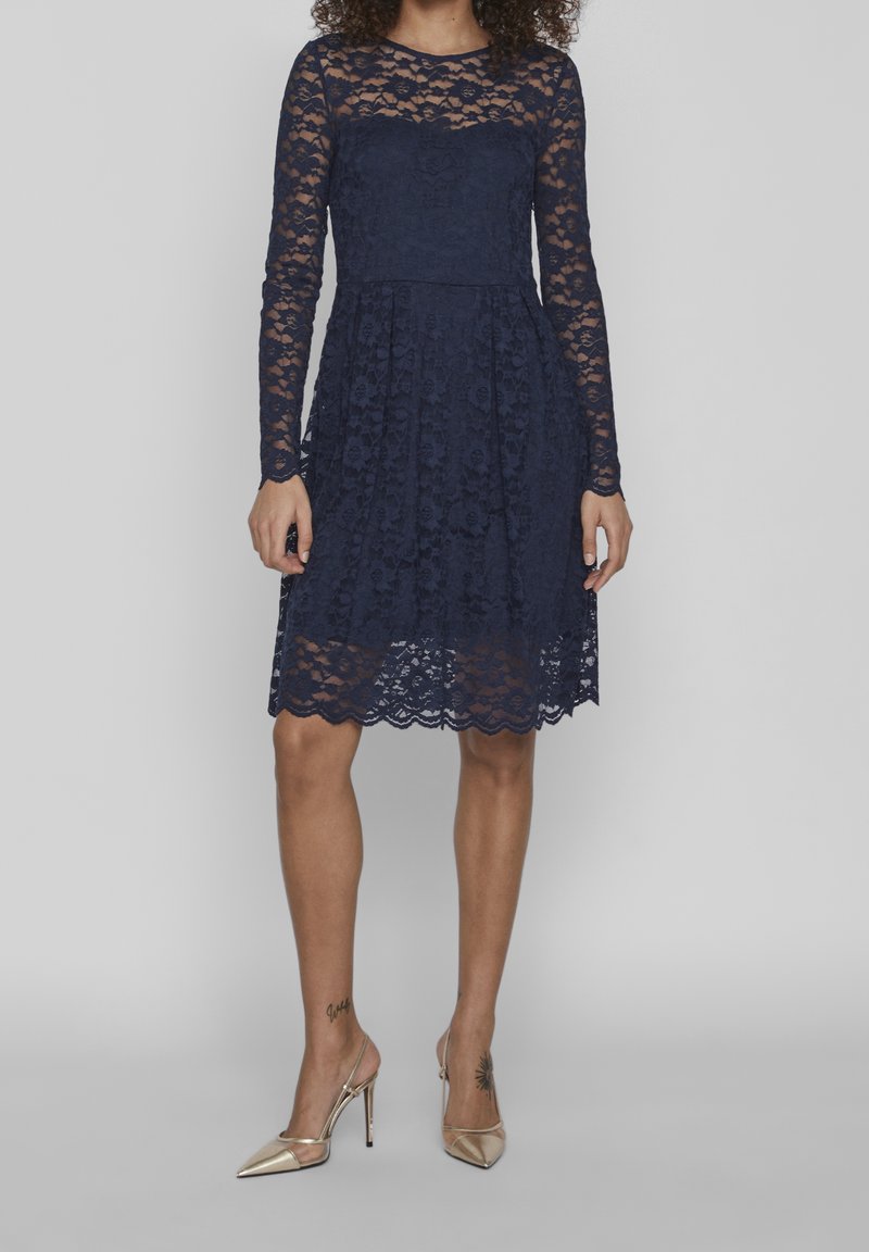 Femme portant une robe en dentelle bleu marine jusqu'aux genoux, à manches longues, et des chaussures à talons beiges, posant devant un fond uni.