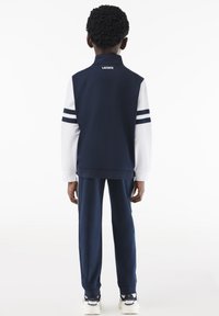 Granatowy dres z białymi długimi rękawami, z trzema poziomymi granatowymi paskami na każdym rękawie i logo Lacoste na kołnierzu.