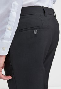 Crne krojene hlače s glatkom teksturom, s stražnjim džepom s detaljem gumba, uparene s bijelom košuljom.