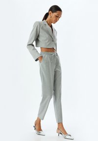 Hellgrauer, taillierter Blazer mit einem einzigen Knopf, kombiniert mit hellgrauen Jogginghosen mit Kordelzug. Silberne Slingback-Schuhe mit spitzem Zehenbereich und Schleifendetail.