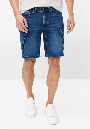Mand iført mørkeblå denimgånger cargo shorts og hvide sneakers, stående med hænderne afslappet ved siderne.