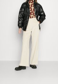 Czarna kurtka puchowa o połyskującej fakturze, brązowy golf z wzorem, kremowe szerokie spodnie sztruksowe oraz masywne czarne buty na platformie.