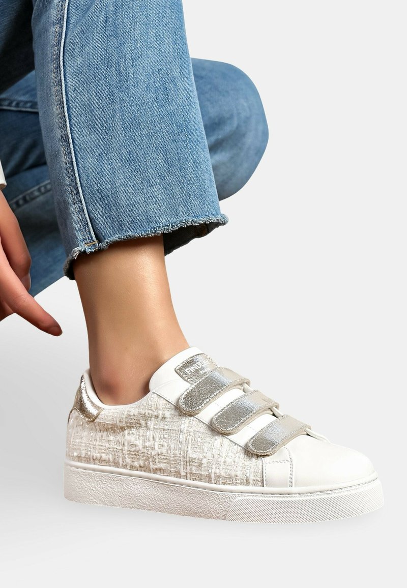 Baskets blanches avec accent beige texturé, bandes velcro argentées, bout rond et semelle en caoutchouc. Design décontracté associé à un jean déchiré.