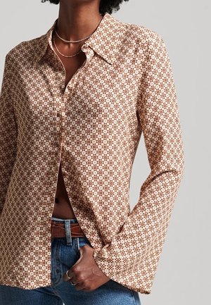 Femme portant une chemise beige à motifs boutonnée avec manches longues, déboutonnée en bas, jean bleu et colliers argentés superposés.