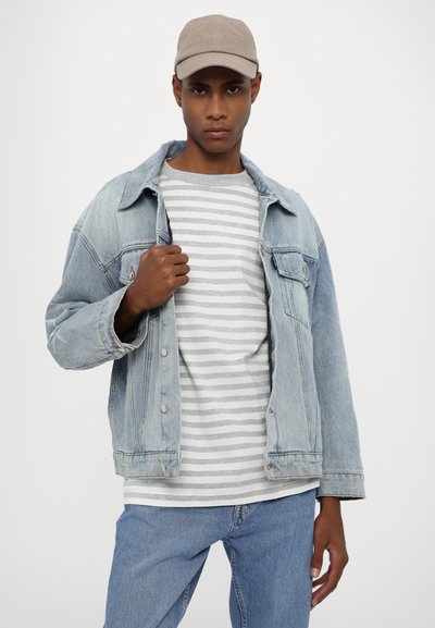 Light blue denim jacket with button closure and wide sleeves, layered over a gray and white striped shirt, paired with blue jeans and a beige cap.