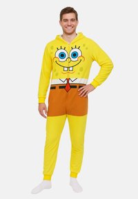 Tuta intera in pile gialla con un design di personaggio dei cartoni animati, grandi occhi blu, una bocca sorridente e pantaloni marroni, con polsini elastici.