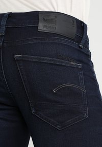 Närbild av bakfickan på mörkblå denimjeans med G-Star RAW-logotyp och böjd sömsdetalj på fickan.