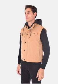 Gilet senza maniche color beige con texture orizzontale, due tasche sul petto e un cappuccio nero, abbinato a una camicia nera a maniche lunghe.