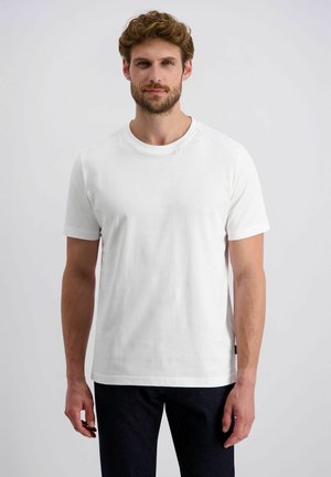 Mann mit Bart und lockigem Haar, der ein schlichtes weißes T-Shirt mit kurzen Ärmeln und dunkle Hosen trägt, steht vor einem schlichten hellen Hintergrund.