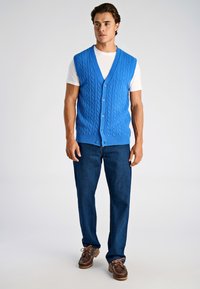 Blå stickad väst med flätmönster, knäppning framtill, bärs över en vit T-shirt. Kombineras med mörkblå jeans och bruna skor.