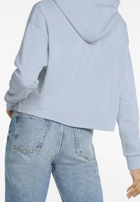 Persoon in een lichtblauwe cropped hoodie en hoge, vervaagde blauwe jeans, weergegeven van achteren met de rechterhand iets uitgestoken.