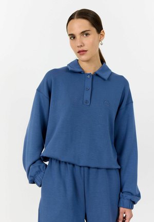 Pullover in cotone blu con collo a camicia, apertura frontale con tre bottoni e vita elastica. Presenta maniche lunghe e un logo discreto.