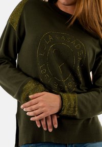 Felpa verde con scollo tondo, caratterizzata da dettagli in strass sulle spalle e un design con logo al centro. Tessuto morbido.