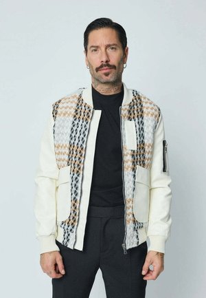 Homme avec moustache et boucles d'oreilles portant une veste blanche à motifs multiples zippée par-dessus une chemise noire, debout devant un fond clair uni.