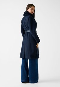 Marineblauwe wollen coat met een nepbont kraag, een riem in de taille en een getailleerde pasvorm, gecombineerd met wijd uitlopende denim broek. Glad textuur, gestructureerd ontwerp.