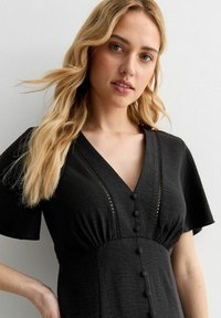 Vestido negro con mangas cortas, escote en V, botones decorativos en el frente y detalles de encaje en el escote. Tejido texturizado.
