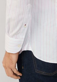 Biela košeľa s tenkými ružovými pruhmi, s gombíkovými manžetami a jemnou textúrou. Viditeľné švy na okrajoch a tónové detaily na rukáve.