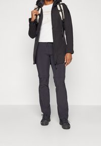 Schwarze wasserdichte Jacke mit Kapuze, Reißverschluss vorne und Taschen; kombiniert mit dunkelgrauen Hosen und schwarzem Schuhwerk. Geeignet für den Outdoor-Einsatz.