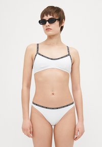 Completo bikini bianco con spalline regolabili. Presenta una banda nera a contrasto con logo sui bordi. Design minimalista, texture liscia.