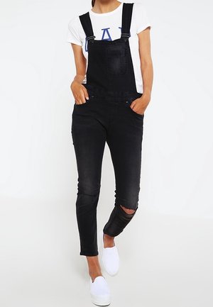 Černé denimové kalhoty na kšandy s nastavitelnými popruhy, přední kapsou a roztrhanými koleny. Kombinované s bílým tričkem a bílými nazouvacími botami.