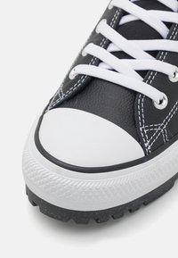 Zapatilla de cuero negro con puntera de goma blanca, costuras blancas y cordones. Presenta una suela texturizada con un prominente dibujo para un mejor agarre.