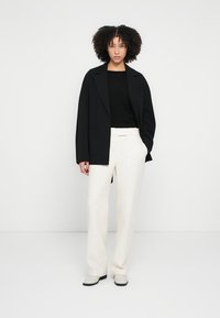 Sacou supradimensionat negru peste un top negru, asortat cu pantaloni largi, texturati, de culoare deschisă. Pantofii alb-negru completează ținuta.