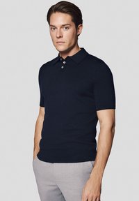 Granatowa koszulka polo z krótkim rękawem, klasycznym kołnierzykiem, trzema guzikami oraz miękką, gładką strukturą. Połączona z jasnoszarymi spodniami.