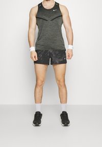 Débardeur de sport noir avec motif texturé et accents en mesh, associé à un short noir à imprimé camouflage. Des baskets noires complètent le look.