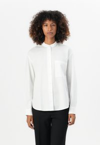 Camicia bianca a maniche lunghe con colletto alla coreana, chiusura a bottoni e taschino sul lato sinistro del petto. Tessuto morbido e vestibilità comoda, abbinata a pantaloni neri.